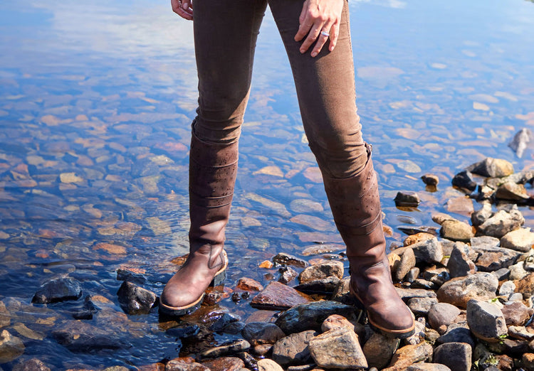 On model image of woman standing in the banks of a river while wearing a pair of Dubarry Roundstone Country Boots, old rum coloured knee high heeled boots with full length zip and leather strap.