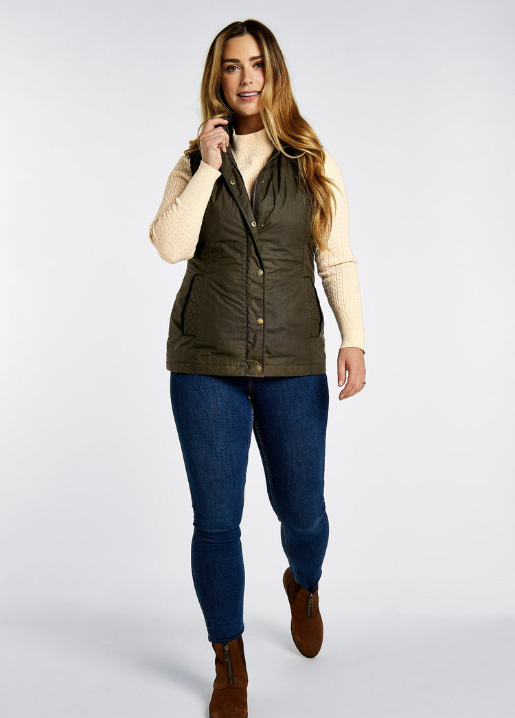 A female model wearing a Dubarry Pakenham wax gilet jacket, ginger brown colour with buttons and zip. The model is also wearing a dark jumper underneath, blue jeans and boots